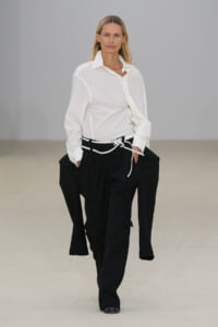 Runway model walking in a crisp white button-up shirt and oversized black trousers with a white belt.