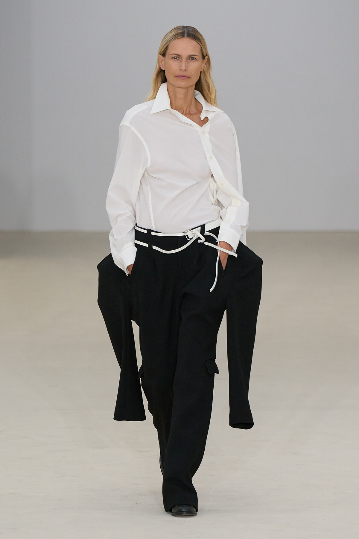Runway model walking in a crisp white button-up shirt and oversized black trousers with a white belt.