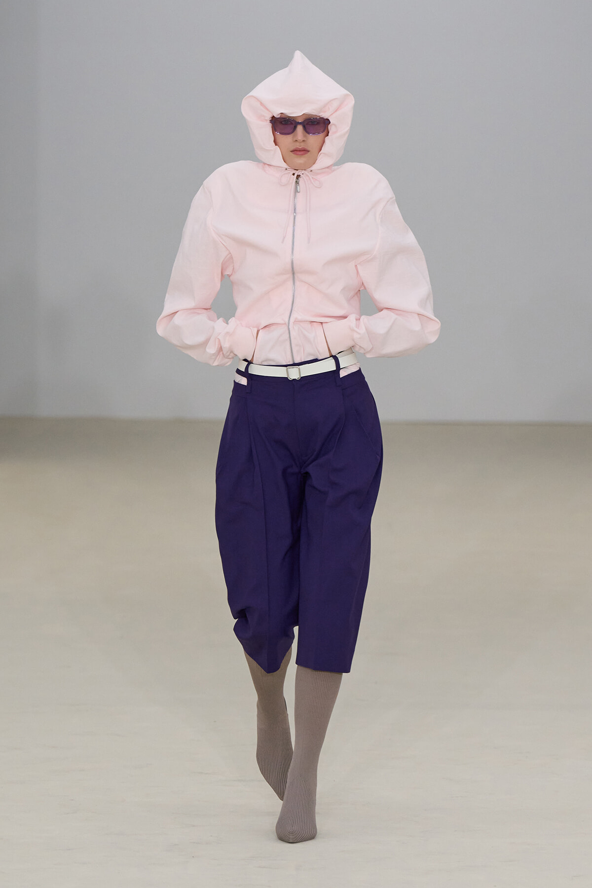 Model on a runway wearing a light pink oversized hooded jacket with zipper, tucked-in white belt, and dark purple wide-leg pants; taupe socks and purple sunglasses add a futuristic vibe.