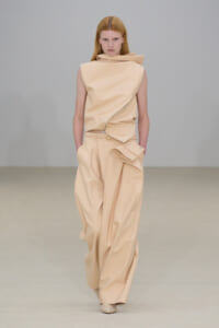 Female model walking the runway in a beige sculptural sleeveless top and wide-leg pants with a wrapped detail. Professional fashion shoot alt text adds the necessary details for screen readers without visual cues.