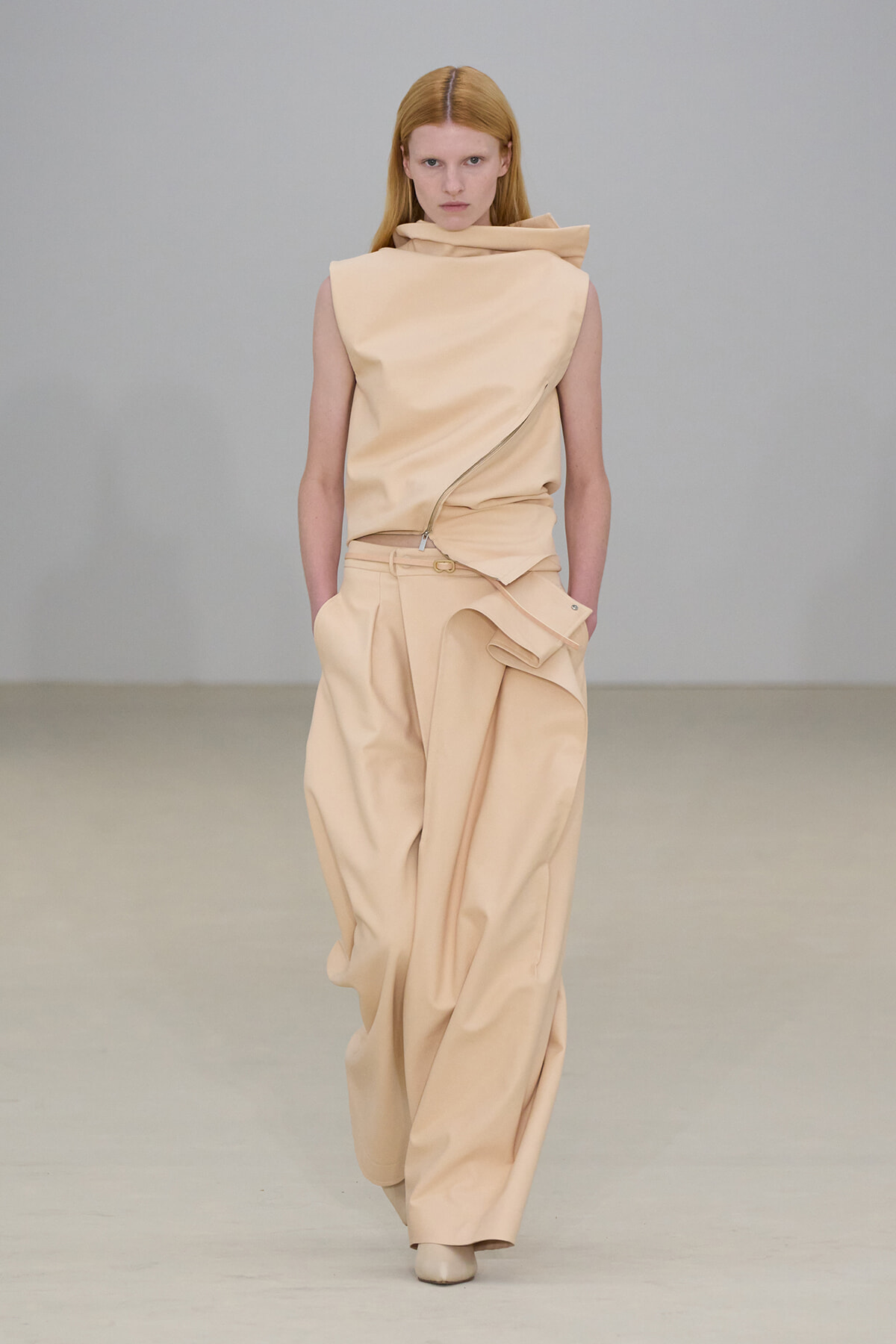 Female model walking the runway in a beige sculptural sleeveless top and wide-leg pants with a wrapped detail. Professional fashion shoot alt text adds the necessary details for screen readers without visual cues.