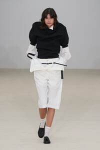 Fashion model on a runway wearing a black puff-sleeve top with white cropped shorts, belt, white socks and black loafers.