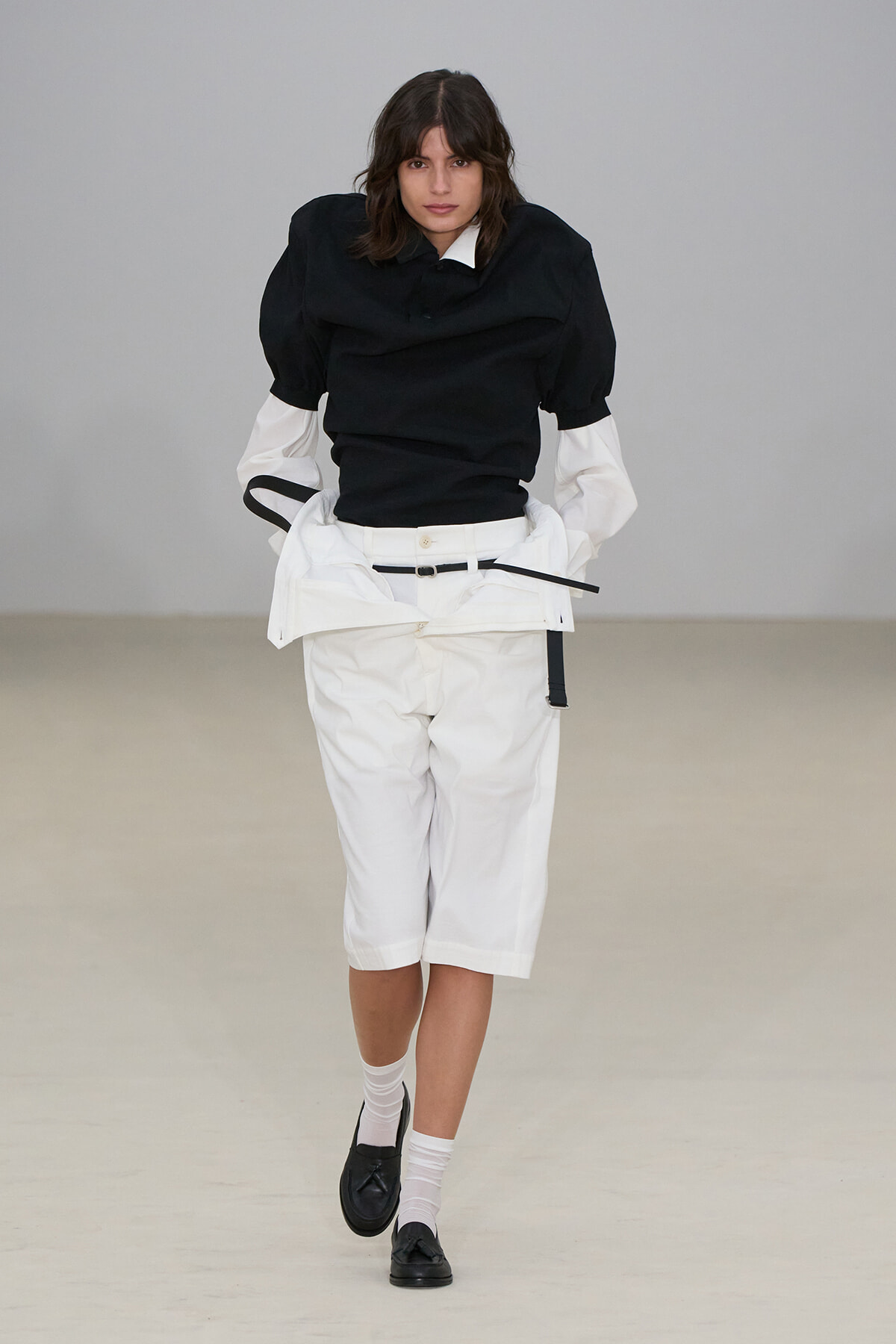 Fashion model on a runway wearing a black puff-sleeve top with white cropped shorts, belt, white socks and black loafers.