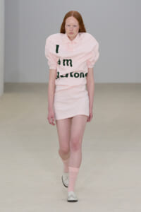 Model walking a runway in a pale pink mini dress with puffed sleeves and large black text across the front.