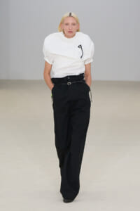 Model walks a minimalist runway in a white puff-sleeve T-shirt tucked into black high-waisted wide-leg trousers with a belt.