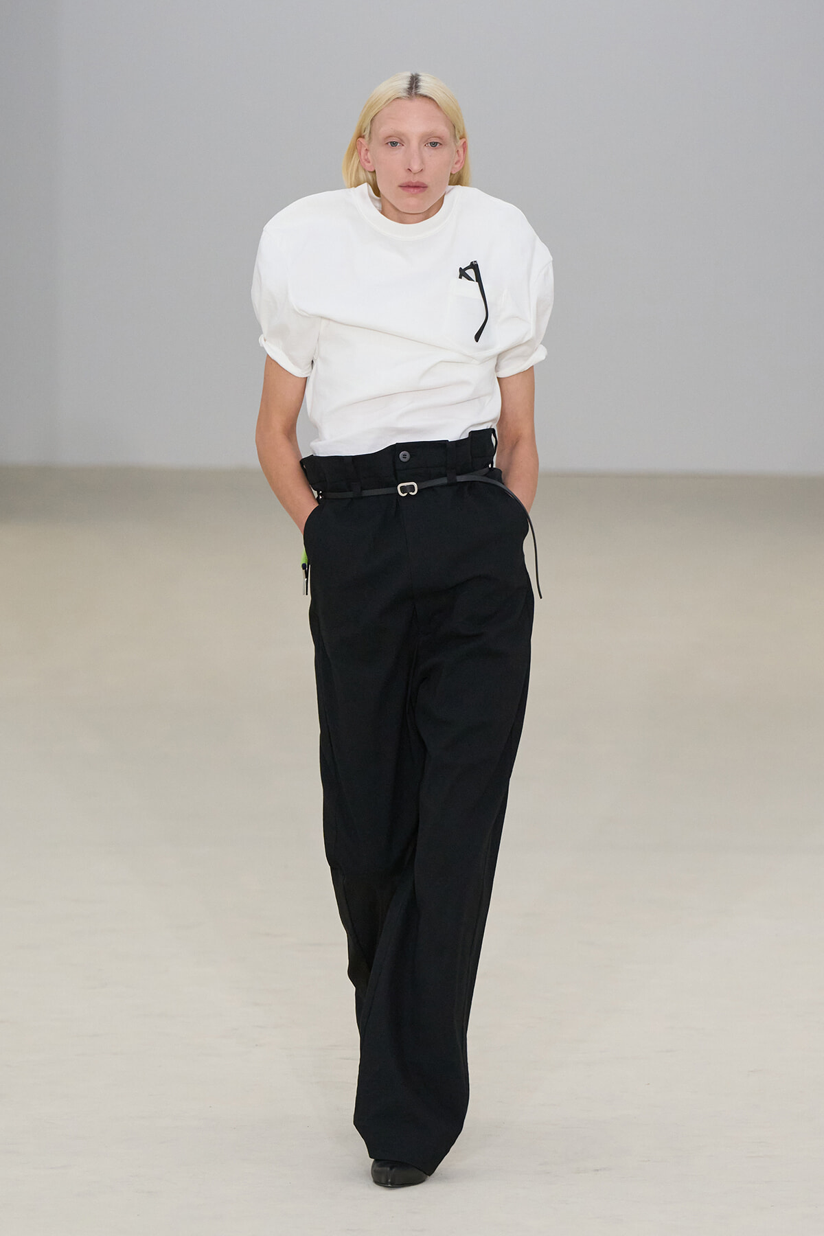 Model walks a minimalist runway in a white puff-sleeve T-shirt tucked into black high-waisted wide-leg trousers with a belt.