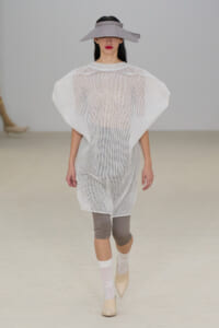 Model walking the runway in a sheer white mesh tunic, taupe wide-brim hat, taupe capri leggings, white socks, and beige heels.