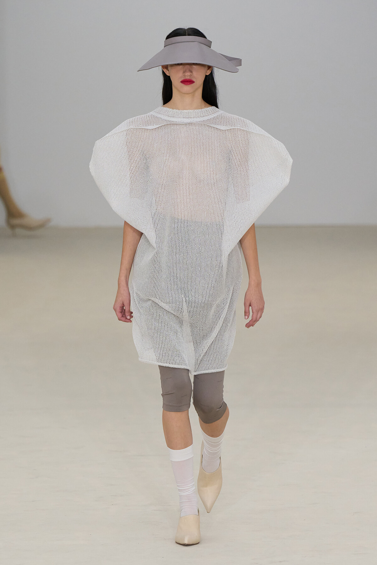 Model walking the runway in a sheer white mesh tunic, taupe wide-brim hat, taupe capri leggings, white socks, and beige heels.