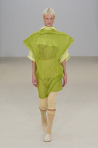 Model walking a runway in a lime-green mesh tunic layered over pale yellow sleeves, with beige leg warmers and boots. Filmed in a minimalist runway setting.