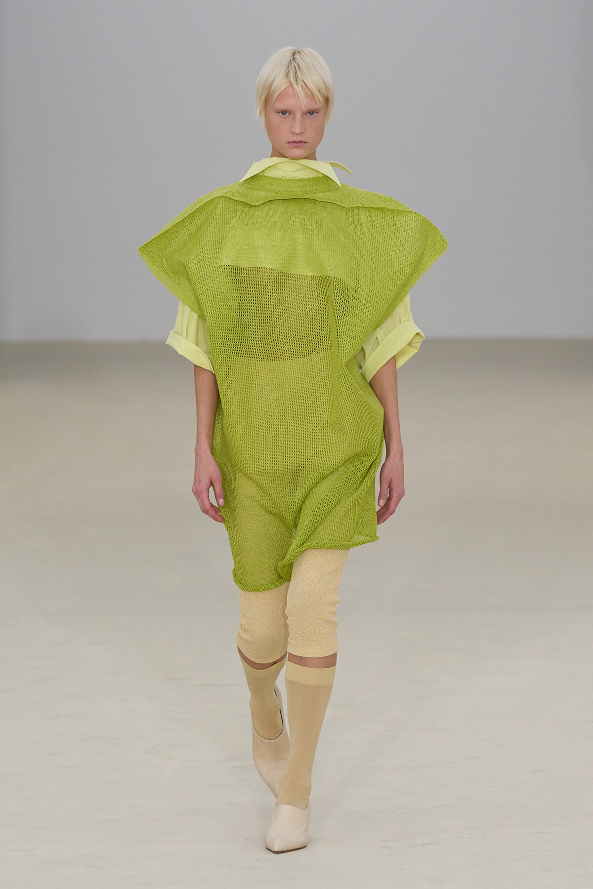 Model walking a runway in a lime-green mesh tunic layered over pale yellow sleeves, with beige leg warmers and boots. Filmed in a minimalist runway setting.