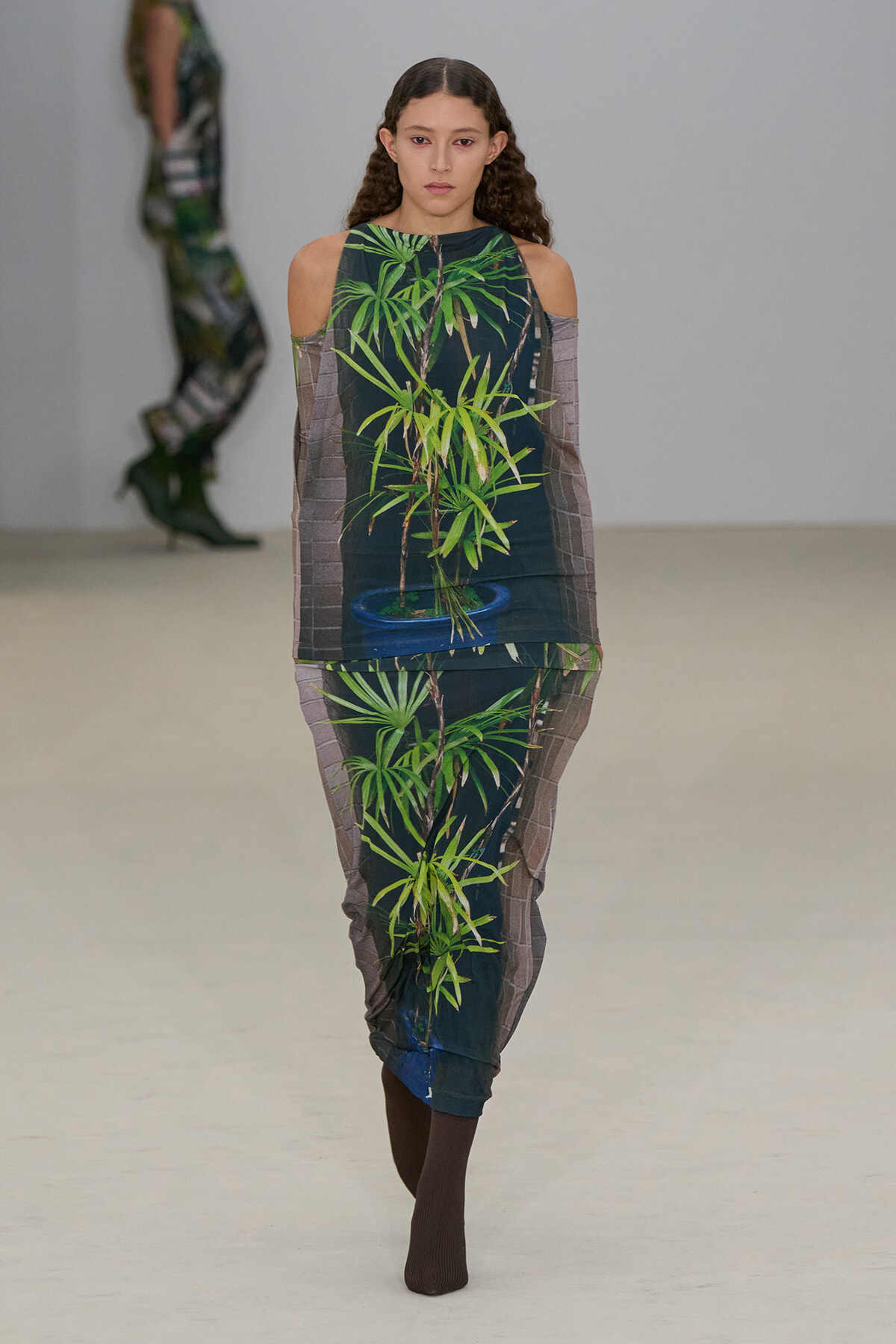 Model on a runway wearing a matching two-piece outfit printed with bright green bamboo plants on a dark background, with sheer brown long sleeves and shoulder cutouts.