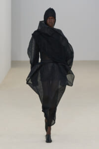 Model walking the runway in a black, oversized mesh ensemble with hooded headpiece and dramatic draping.
