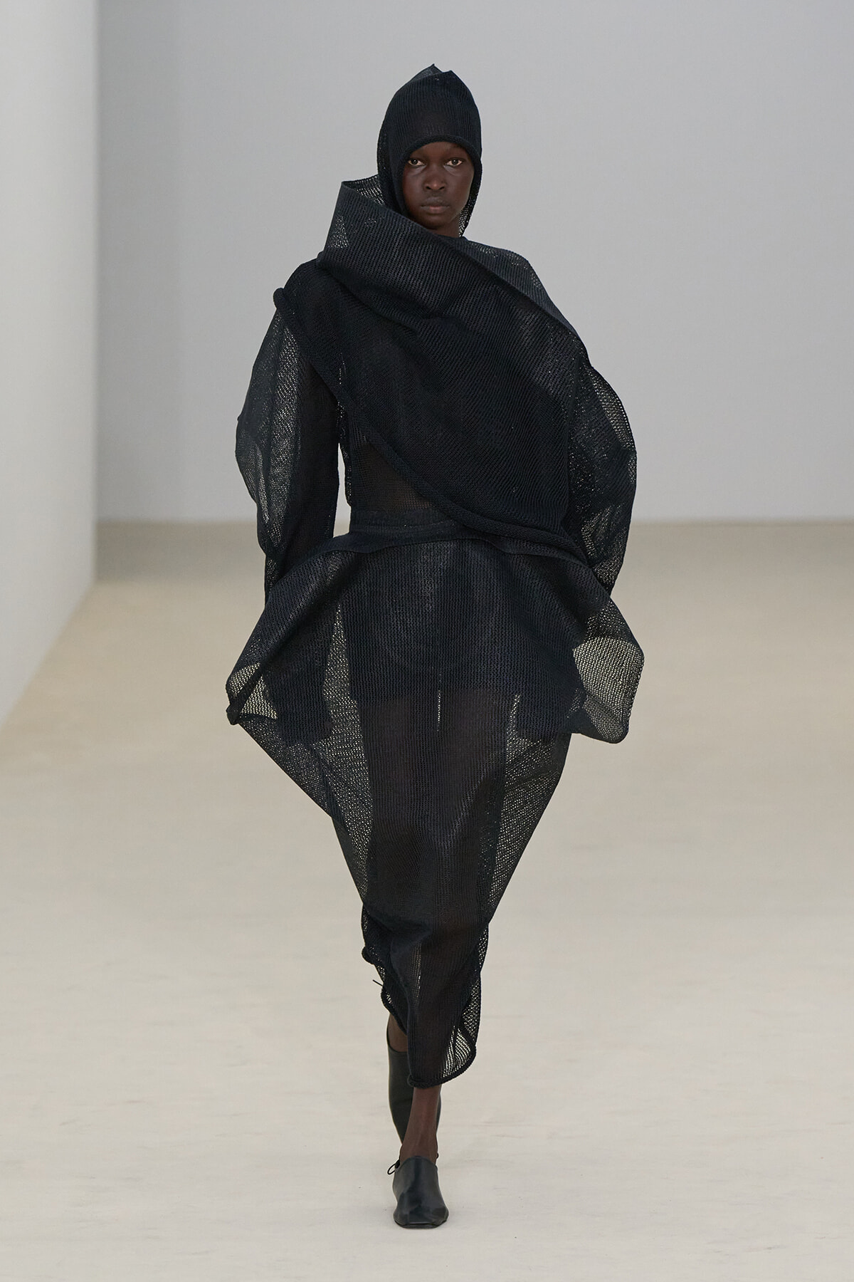 Model walking the runway in a black, oversized mesh ensemble with hooded headpiece and dramatic draping.