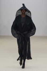 Model walking in a black sheer, hooded robe with a tied belt on a blank runway.