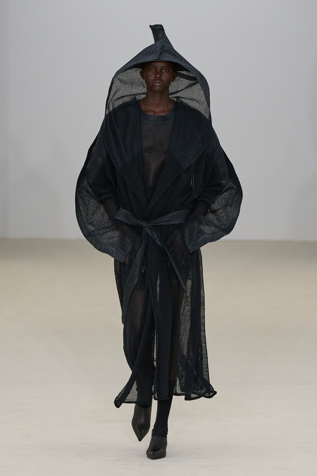 Model walking in a black sheer, hooded robe with a tied belt on a blank runway.