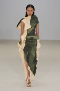 Runway model wearing a color-blocked green and cream draped gown with sculptural, asymmetric panels and white sandals.