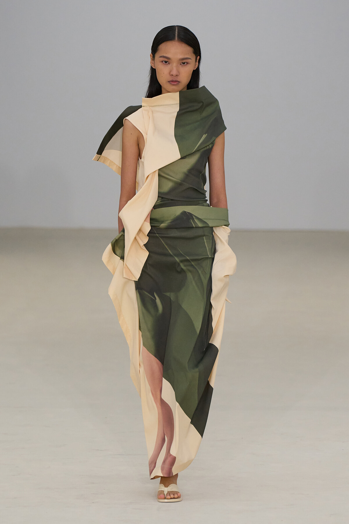 Runway model wearing a color-blocked green and cream draped gown with sculptural, asymmetric panels and white sandals.