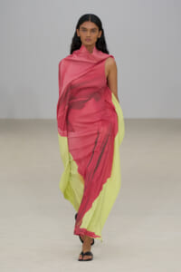 Model walking runway in a pink and lime-green draped gown, one-shoulder design with flowing fabric and black sandals