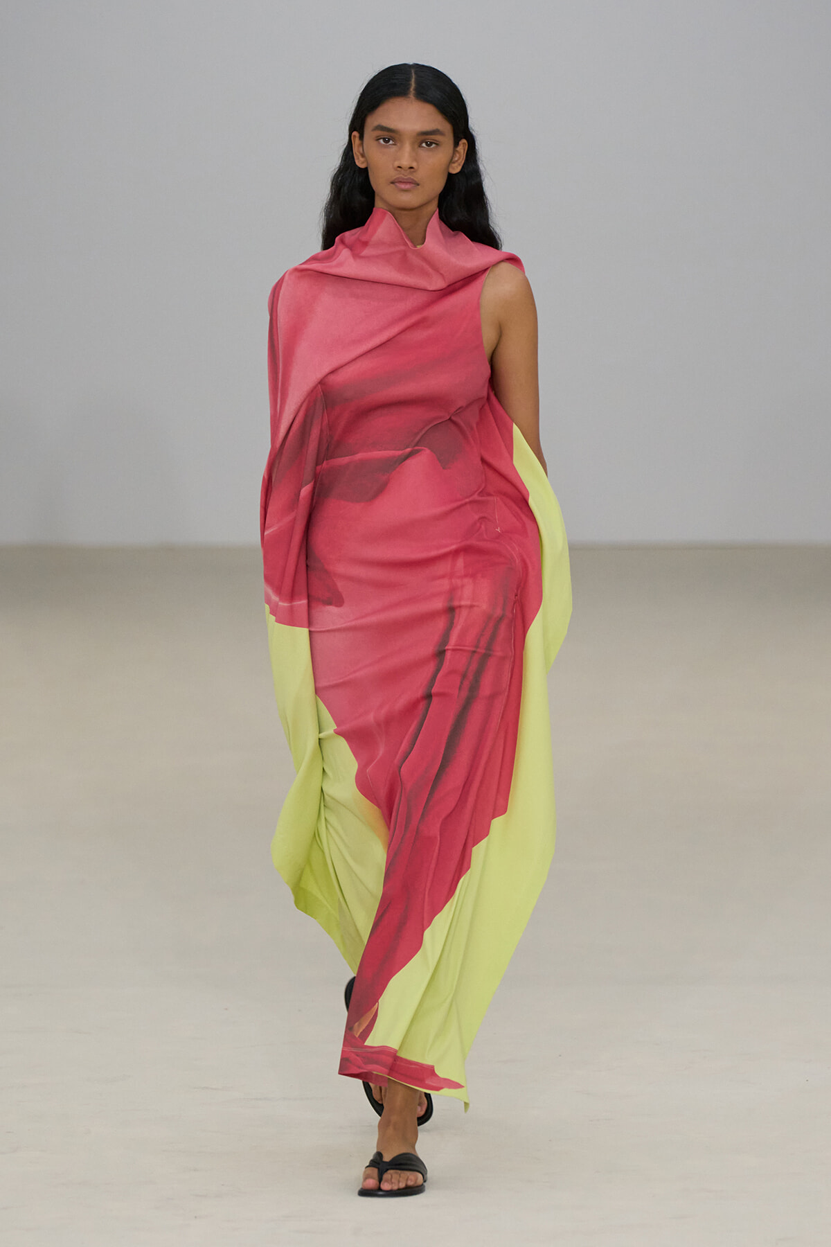 Model walking runway in a pink and lime-green draped gown, one-shoulder design with flowing fabric and black sandals