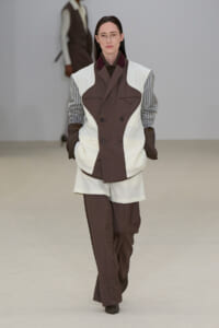 Female model walking a runway in a brown-and-white color-block suit with striped sleeves and brown pants, wearing gloves and glasses.