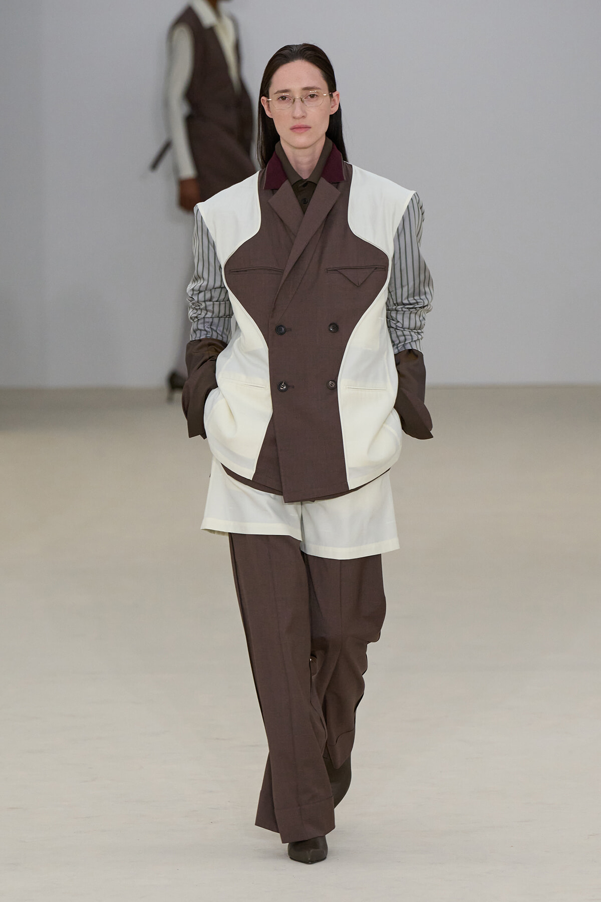 Female model walking a runway in a brown-and-white color-block suit with striped sleeves and brown pants, wearing gloves and glasses.