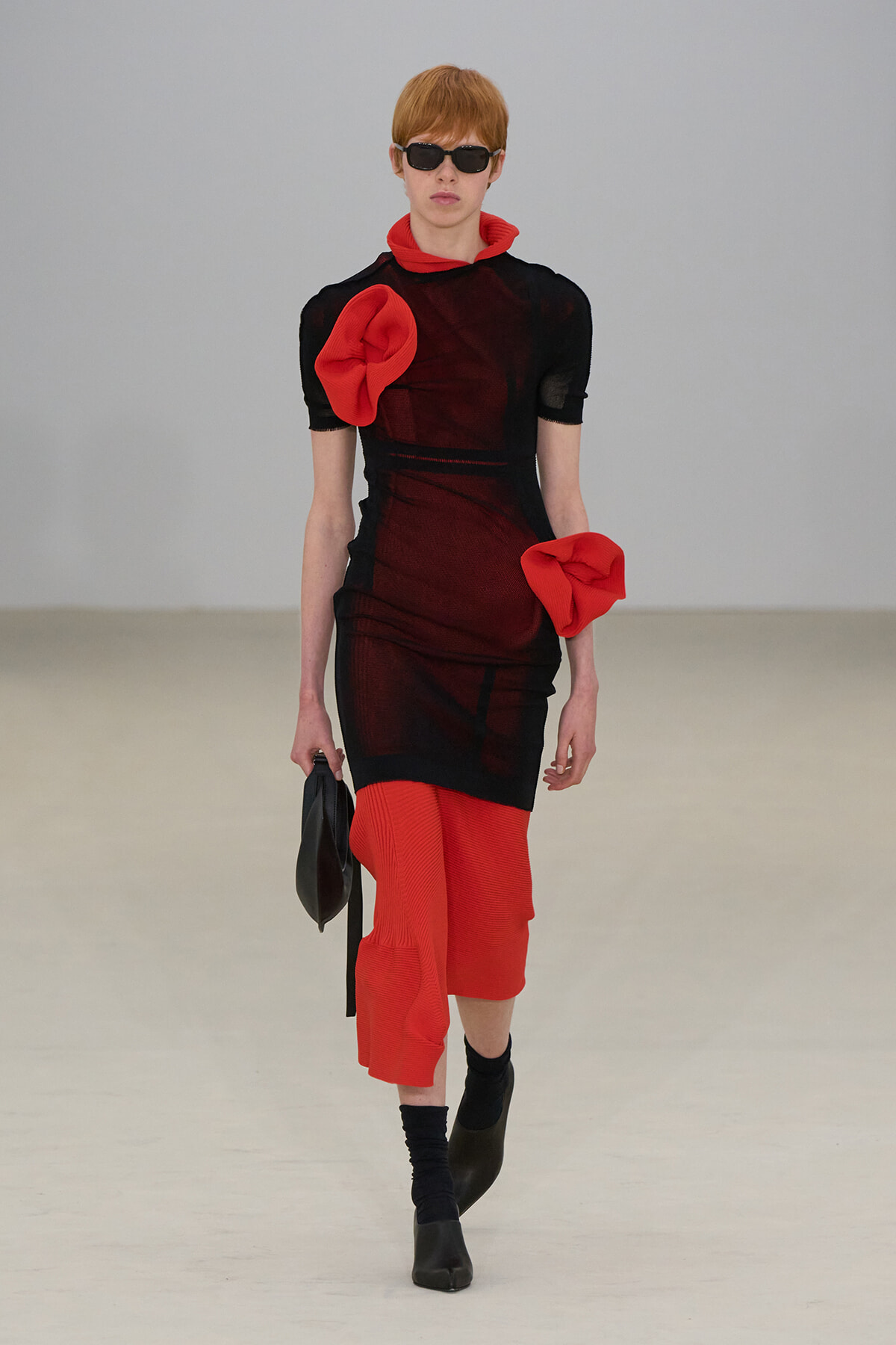 Model wearing a sheer black dress with bold red accents, red scarf at the neck, oversized red bow on the shoulder, and red cropped pants, plus black sunglasses and clutch.