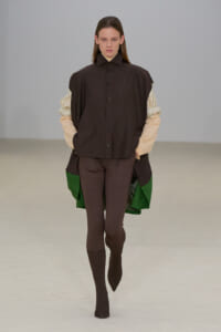 Model on a runway wearing an oversized dark brown button-up over beige striped sleeves, brown leggings, and dark brown socks; green lining shows at the sides.