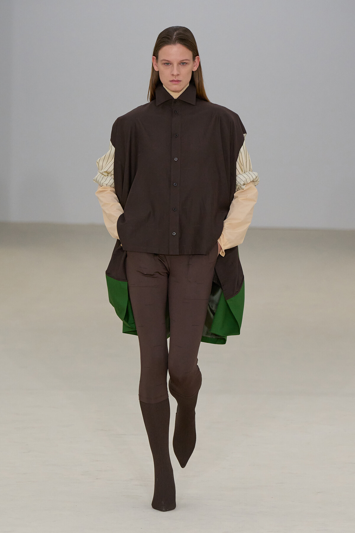Model on a runway wearing an oversized dark brown button-up over beige striped sleeves, brown leggings, and dark brown socks; green lining shows at the sides.