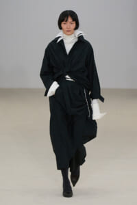 Model walking a runway in a black, oversized layered outfit with a twisted front knot and white collar peeking out, against a neutral backdrop.