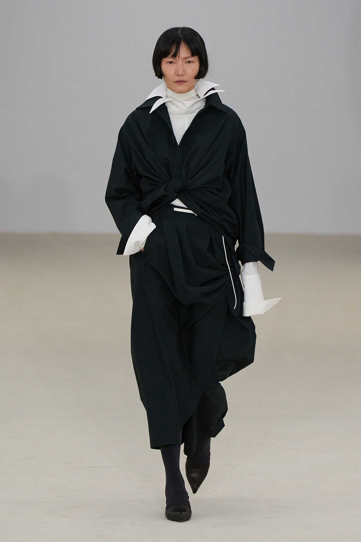 Model walking a runway in a black, oversized layered outfit with a twisted front knot and white collar peeking out, against a neutral backdrop.