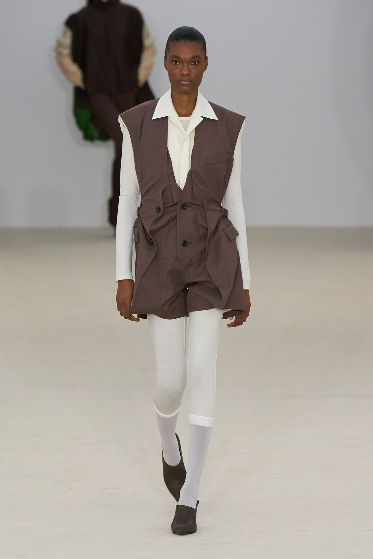 Female model walks a runway in a brown sleeveless blazer over a cream shirt, white leggings, and black shoes.