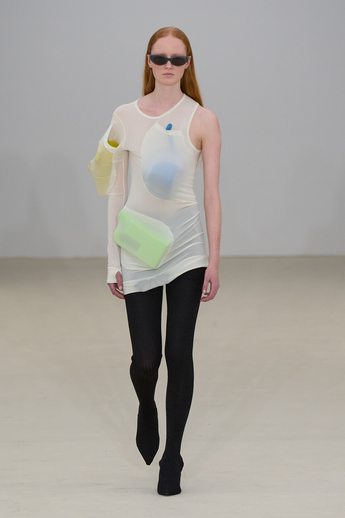 Model walking the runway in a sheer cream top with pastel-colored pockets and black tights, wearing sunglasses.
