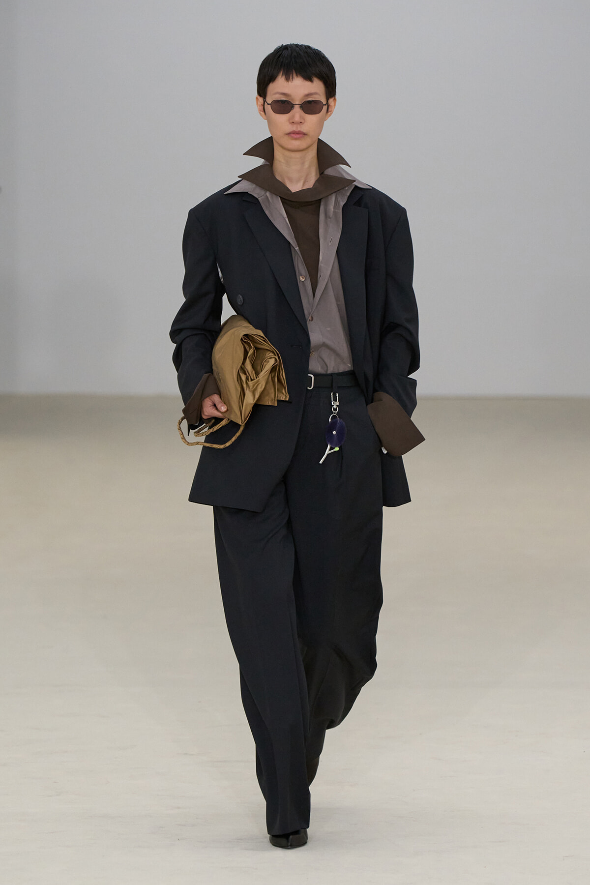 Model walking in a dark suit with brown shirt and oversized collar, holding a tan garment bag and keys on a runway.