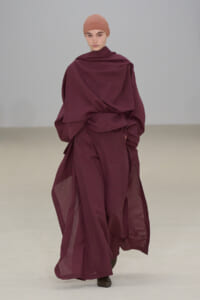 Model walking a runway in a burgundy, layered draped outfit with a matching headscarf.