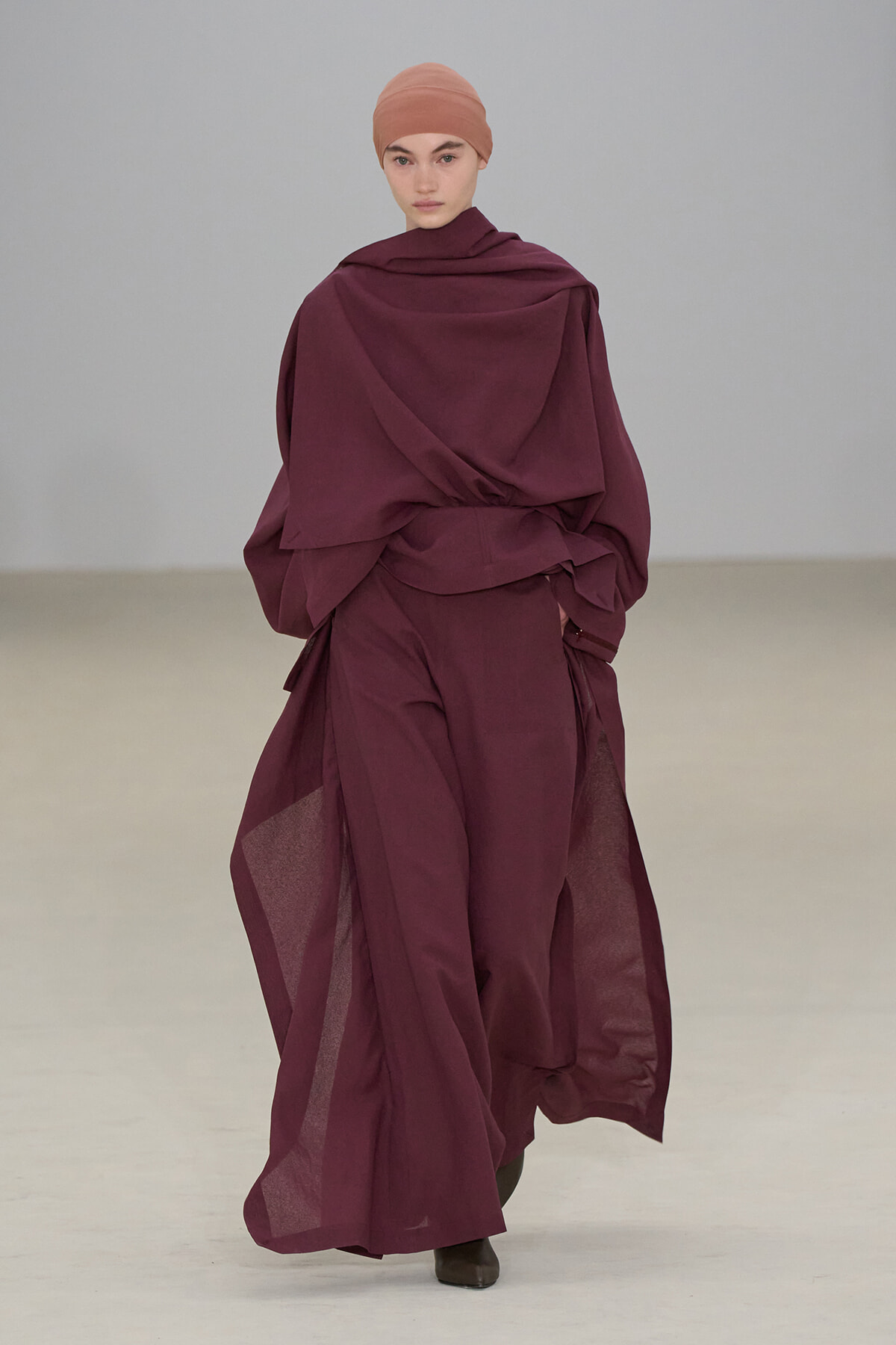 Model walking a runway in a burgundy, layered draped outfit with a matching headscarf.