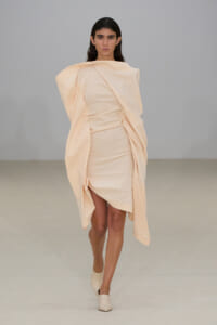 Model walks a runway in a pale peach, draped wrap dress with sculptural volume on the shoulders and sleeves.