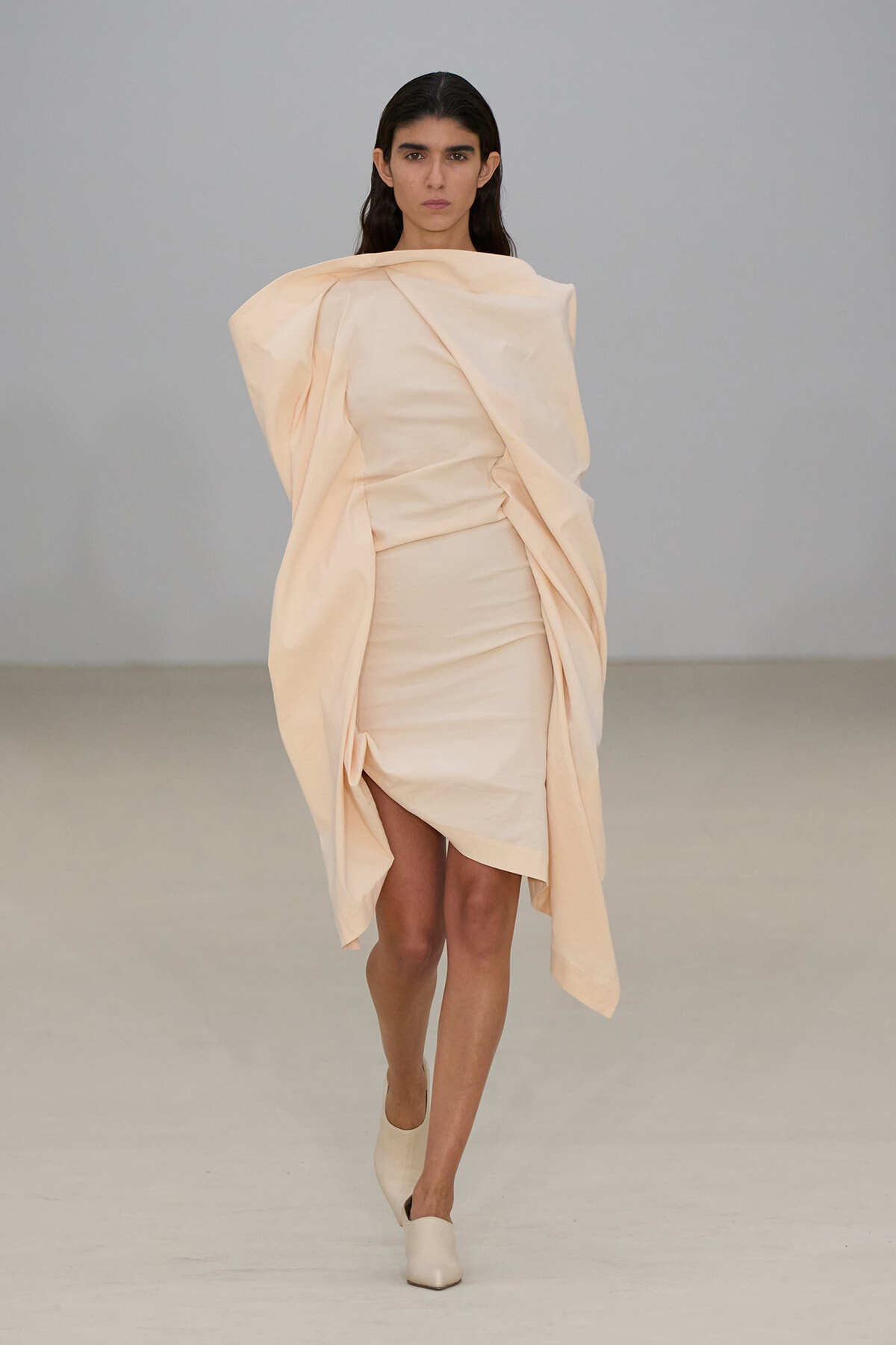 Model walks a runway in a pale peach, draped wrap dress with sculptural volume on the shoulders and sleeves.