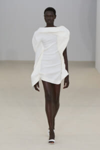 Model walking a runway in a white sculptural mini dress with oversized draped collar and folded details