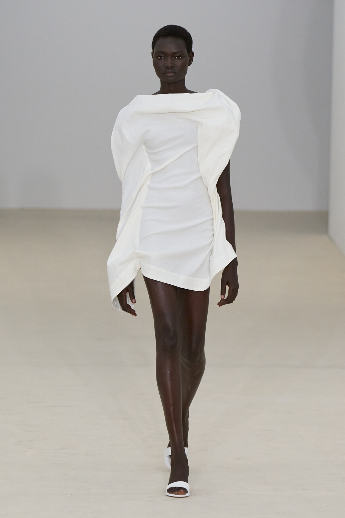 Model walking a runway in a white sculptural mini dress with oversized draped collar and folded details