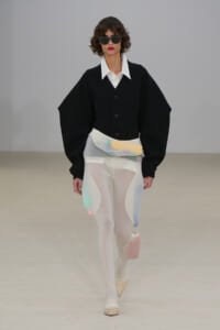Runway model in a black oversized jacket with a white shirt, sheer white tights with pastel color panels, and a pastel waist bag with sunglasses.