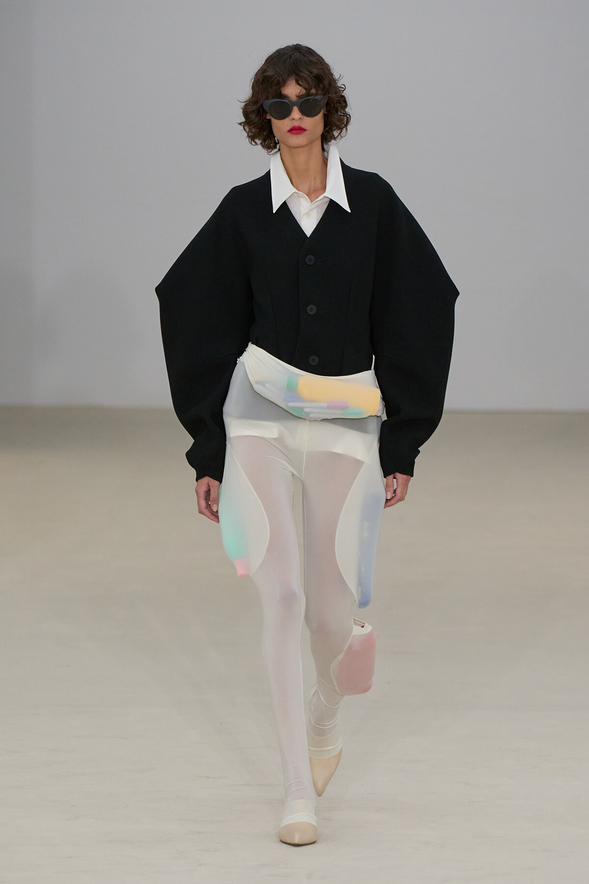 Runway model in a black oversized jacket with a white shirt, sheer white tights with pastel color panels, and a pastel waist bag with sunglasses.