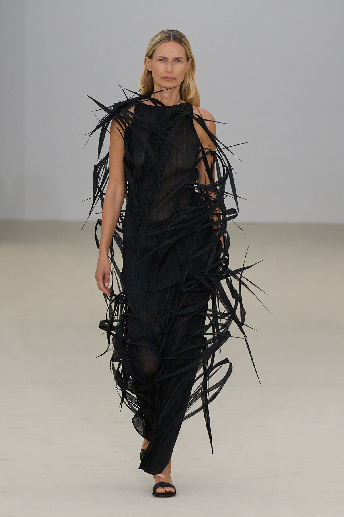Female model wearing a long black sleeveless gown with dramatic jagged fringe strips cascading over the dress on a runway.
