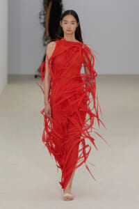 Model walking the runway in a bright red, fringed gown with long, shredded strips cascading around her body.