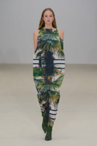 Model walks a tropical print matching top and pants set featuring palm tree motif and green ankle boots.