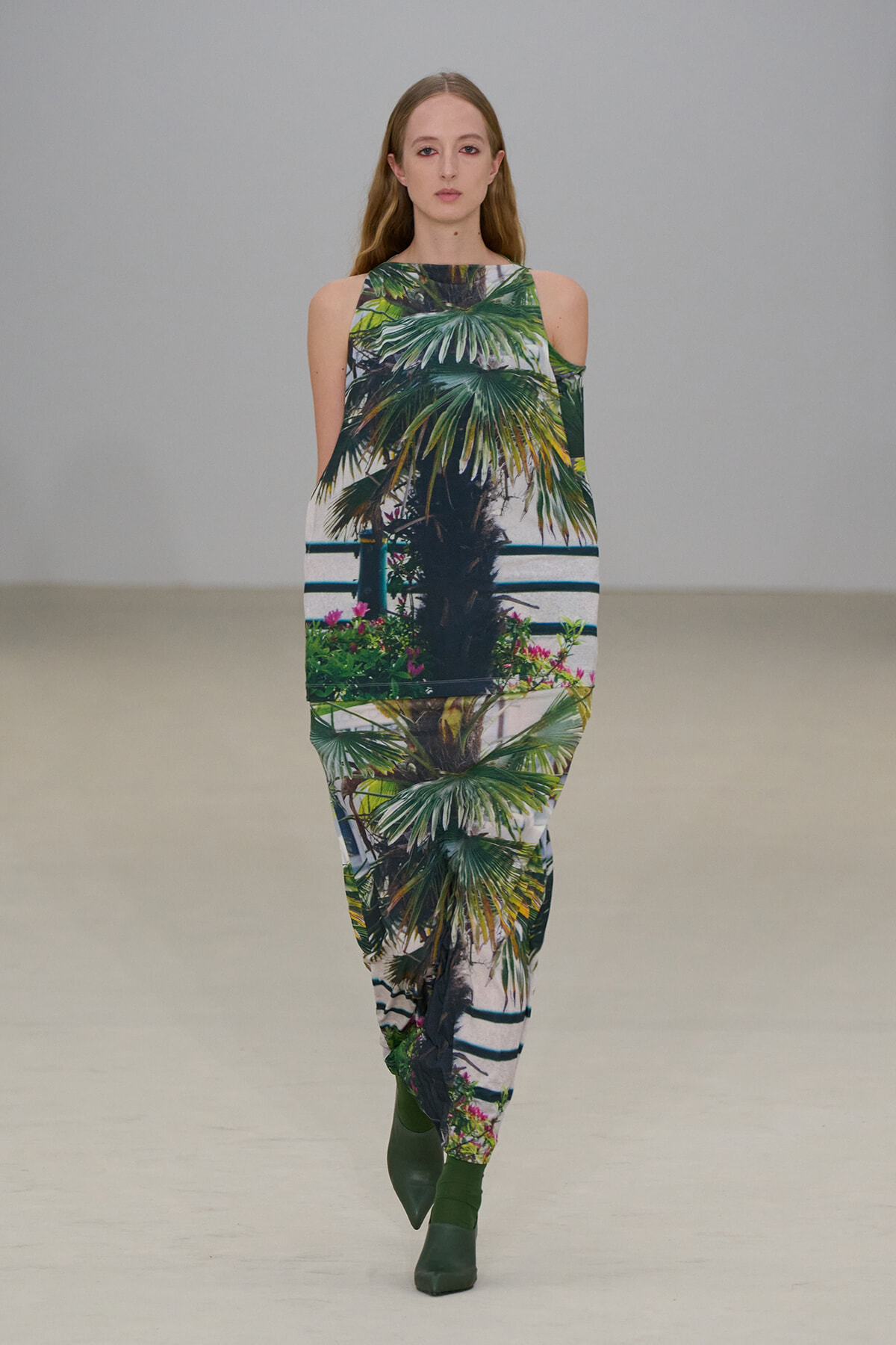 Model walks a tropical print matching top and pants set featuring palm tree motif and green ankle boots.