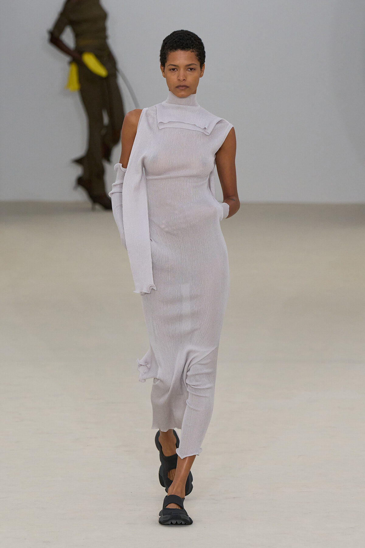 Model walking the runway in a pale gray knit sleeveless turtleneck dress with a draped long sleeve and ruched neckline detail.