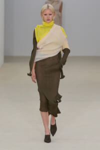 Model walking a runway in a layered, color-blocked outfit: neon yellow top, cream wrap over brown ensemble, and pointed heeled boots.