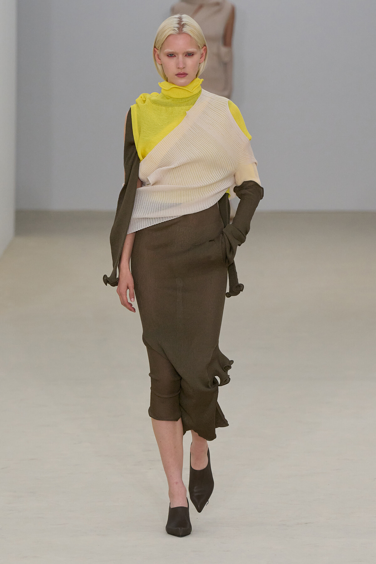 Model walking a runway in a layered, color-blocked outfit: neon yellow top, cream wrap over brown ensemble, and pointed heeled boots.