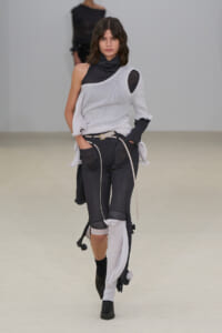 Model walking a runway in a light gray, asymmetrical knit top over a black turtleneck, paired with dark, patchwork-style pants and black ankle boots.