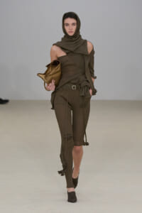 Model walking a runway in an olive-brown knit outfit with a hood, one-shoulder top, and belted pants, carrying a bronze clutch.